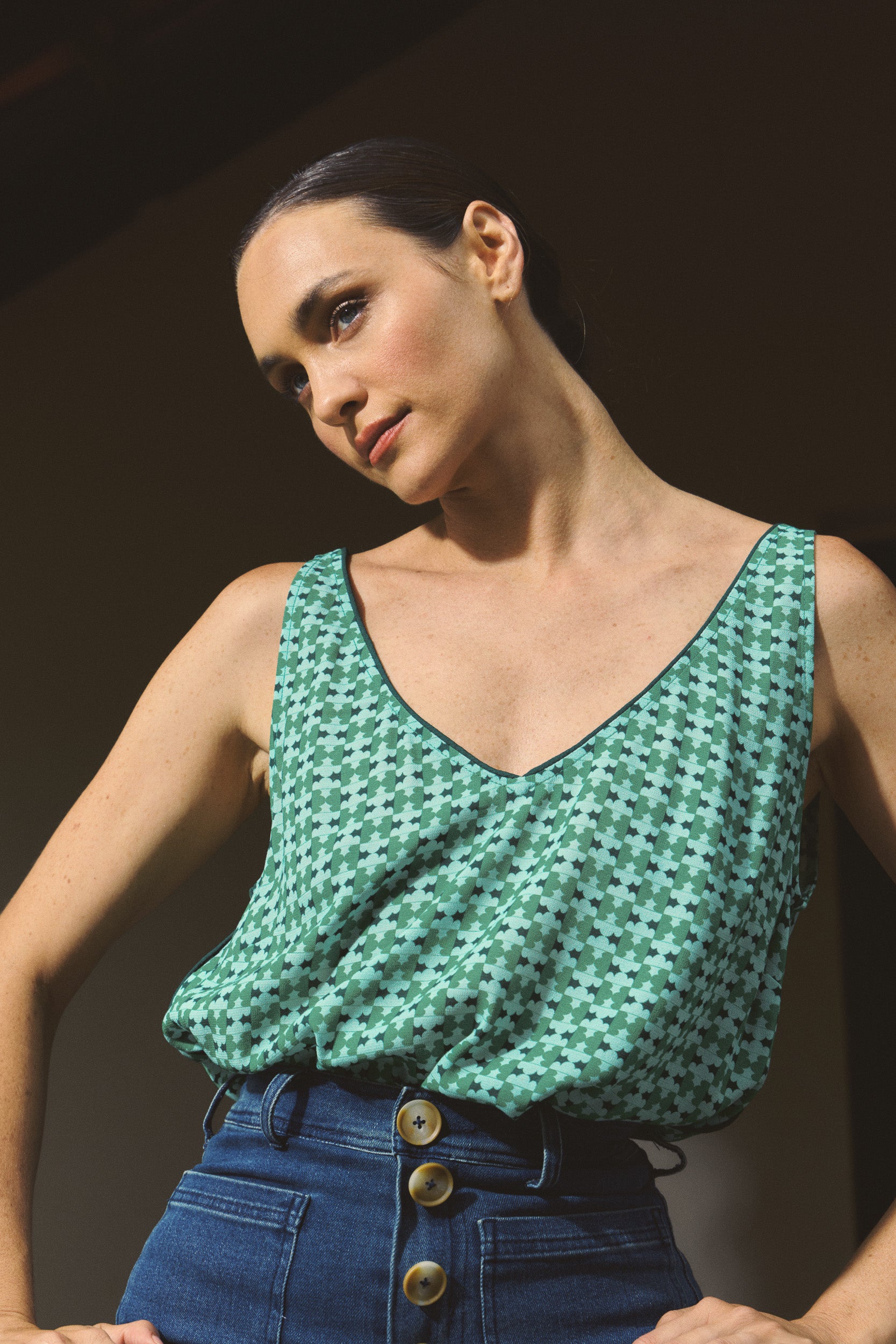 Woman wearing a green patterned top and blue high-waisted jeans against a dark background