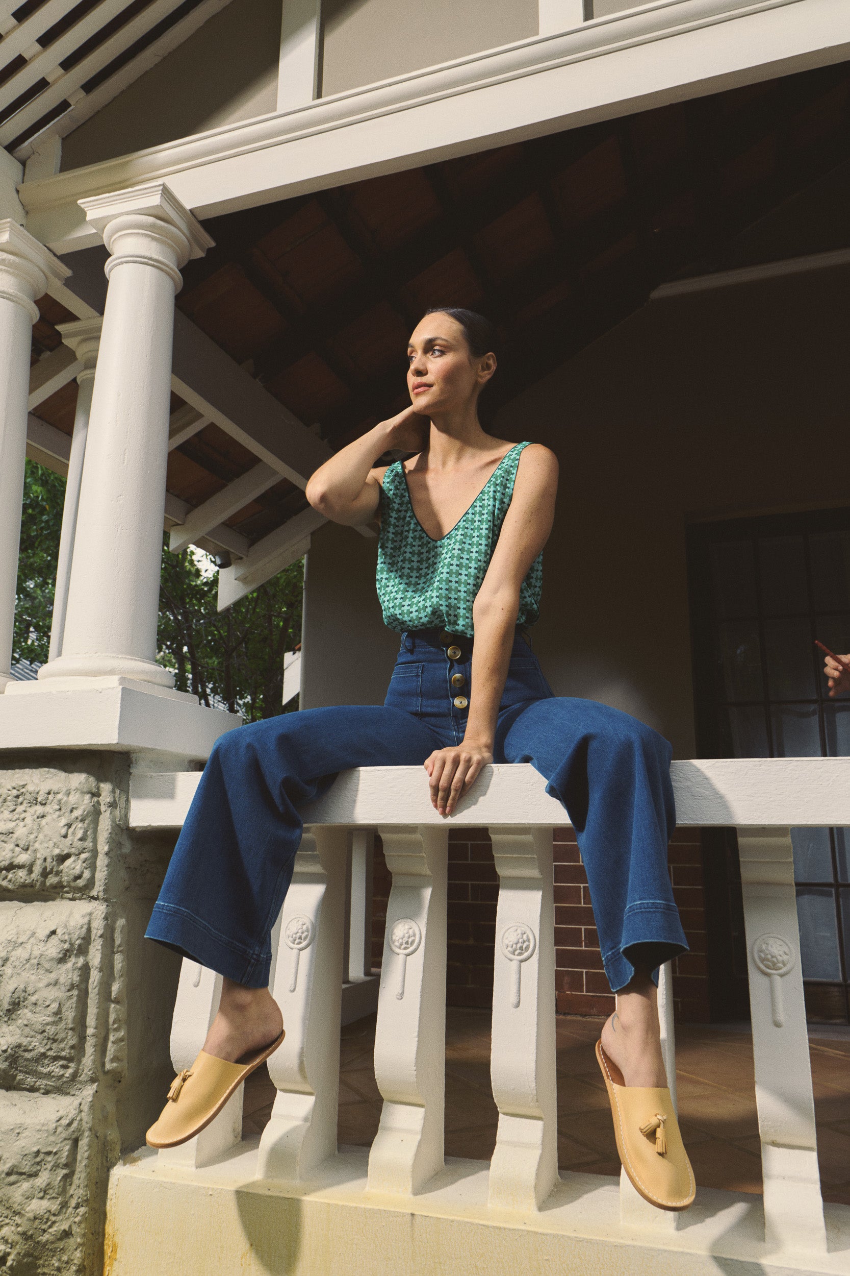 Woman sitting on a white railing outdoors, wearing a green patterned tank top and blue high-waisted jeans.